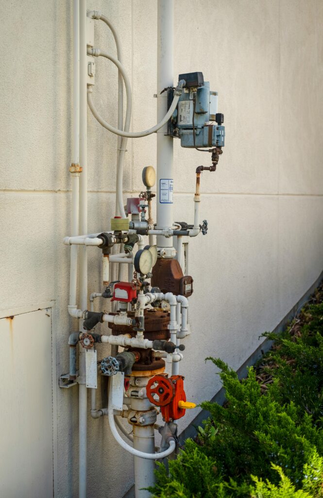 Detailed view of industrial pipes and meters against a concrete wall with green plants below.
