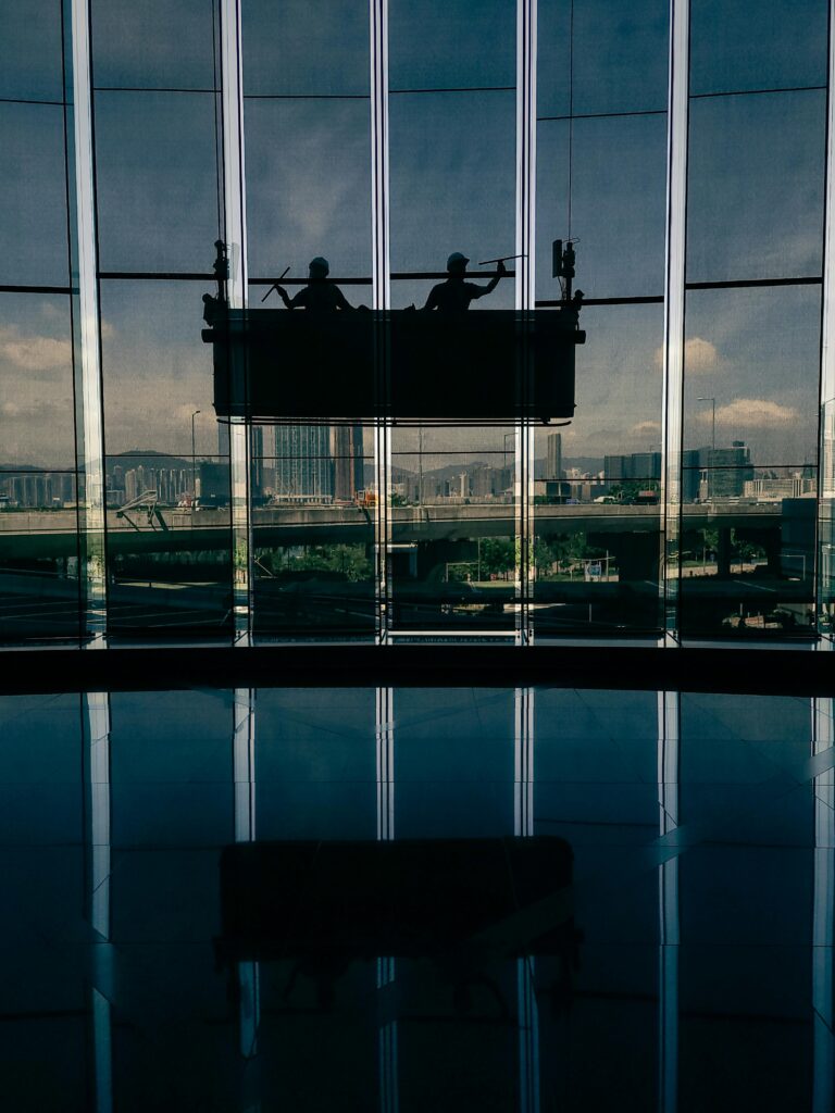 Silhouettes of two window cleaners working on a high-rise building with city skyline reflection in glass windows.